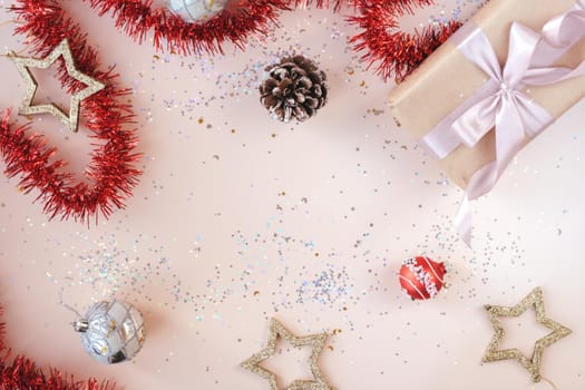 Christmas or New Year festive background with space for text. Present in kraft paper with pink ribbon on soft beige background with glitter, stars, balls, confetti, tinsel. Xmas composition. Flat lay