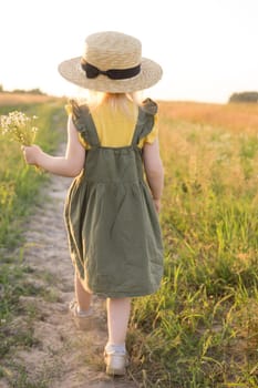 A little blonde girl in a straw hat walks in a field with a bouquet of daisies. The concept of walking in nature, freedom and an eco-friendly lifestyle.