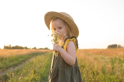 A little blonde girl in a straw hat walks in a field with a bouquet of daisies. The concept of walking in nature, freedom and an eco-friendly lifestyle.