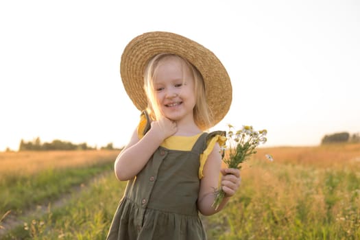 A little blonde girl in a straw hat walks in a field with a bouquet of daisies. The concept of walking in nature, freedom and an eco-friendly lifestyle.