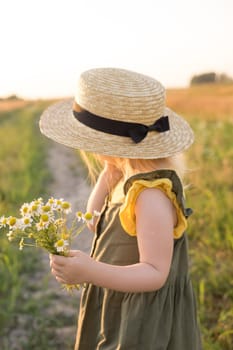 A little blonde girl in a straw hat walks in a field with a bouquet of daisies. The concept of walking in nature, freedom and an eco-friendly lifestyle.