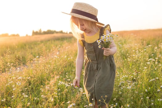 A little blonde girl in a straw hat walks in a field with a bouquet of daisies. The concept of walking in nature, freedom and an eco-friendly lifestyle.