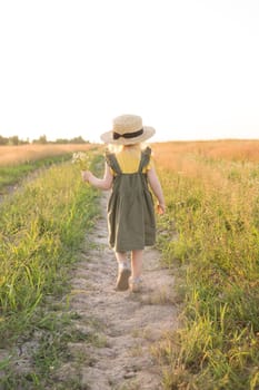 A little blonde girl in a straw hat walks in a field with a bouquet of daisies. The concept of walking in nature, freedom and an eco-friendly lifestyle.