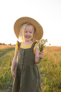 A little blonde girl in a straw hat walks in a field with a bouquet of daisies. The concept of walking in nature, freedom and an eco-friendly lifestyle.