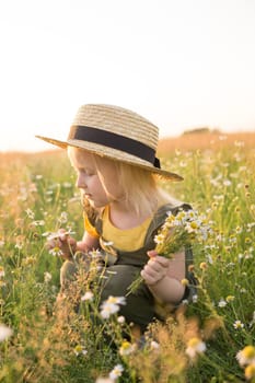 A little blonde girl in a straw hat walks in a field with a bouquet of daisies. The concept of walking in nature, freedom and an eco-friendly lifestyle.