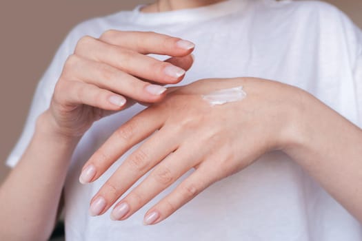 Female hands with natural pink manicure applying moisturizing cream or lotion on beige background. Bodycare, skin protection concept. Woman in white uses beauty product. Spa procedure closeup.