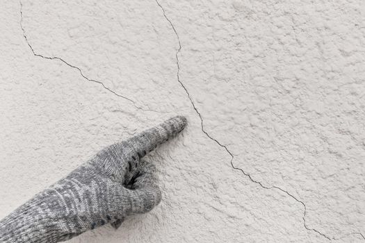 Hand of industrial worker in construction glove points to cracks on white plaster wall texture background.