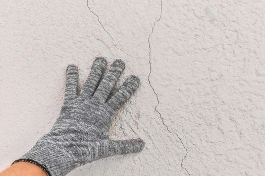 Hand of a working man in a construction glove examines cracks on a white wall background. Repair concept.