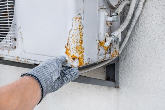 Hand of industrial worker in construction glove touching rust on old air conditioner against white wall background.