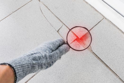 The hand of an industrial worker in a construction glove with a magnifying glass examines the damaged cracked tile background. Renovation concept.