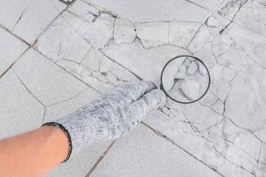 The hand of an industrial worker in a construction glove with a magnifying glass examines the old damaged gray tile background. Renovation concept.