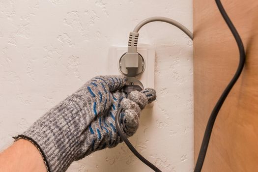 The hand of a male construction worker in protective gloves connects a plug to a double socket against the background of a plaster wall.