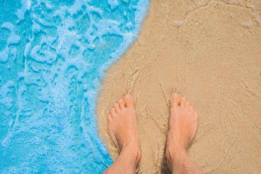 The young man's legs stand on the beach sand and are washed by blue sea water, a view from above.