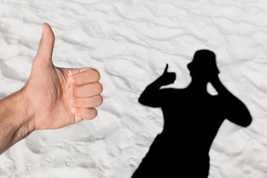 The man's hand shows a cool finger up against the background of his black silhouette shadow on the white beach sand.