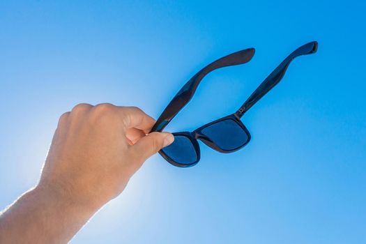 The young man's hand holds sunglasses against the blue sky and sun.