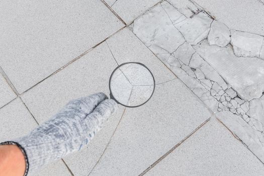 The hand of an industrial worker in a construction glove with a magnifying glass examines the damaged cracked tile background. Renovation concept.