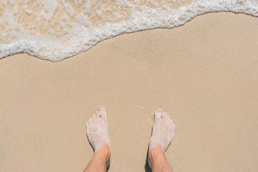 The young man's legs stand on the beach sand and are washed by blue sea water, top view.