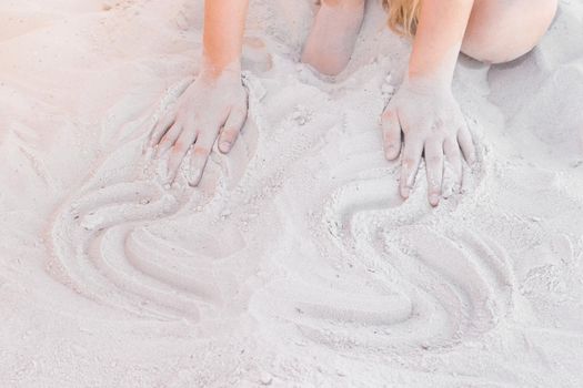 The hands of a young girl draw abstract patterns on a white beach sand background.