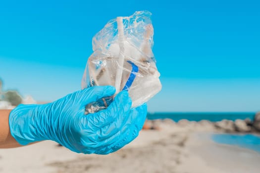 The hands of a man in blue household protective gloves hold a pile of garbage in a bag against the sky and sea beach, close-up.