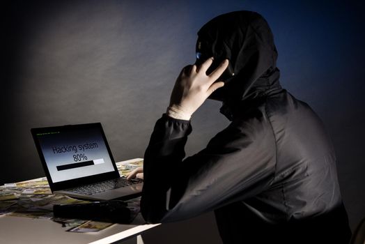 Anonymous hacker in mask sitting sideways uses a laptop and talking on the phone to hack the system in the dark. The concept of hacking database. Money is scattered on the table. Focus on laptop