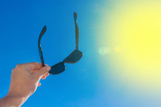 The young man's hand holds sunglasses against the blue sky and sun.