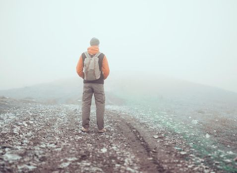 Hiker man with backpack standing on road in the mist, rear view