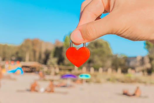 The hand of a man holds a small red heart in the form of a castle close-up against the sea beach with holidaymakers in the resort area. Sign or symbol of love concept.