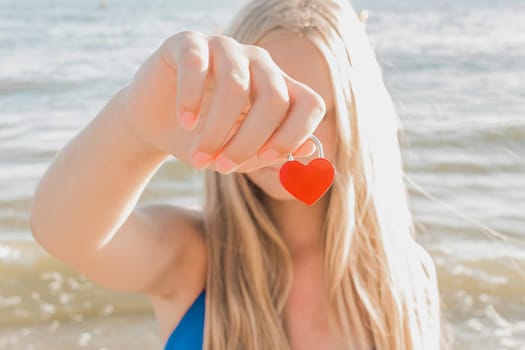 A young blonde girl holds in her hand a small red heart in the form of a castle against the sea. Sign and symbol of love concept.