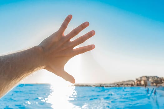 The man's hand is close-up waving against the bright sun, blue sea and horizon line.