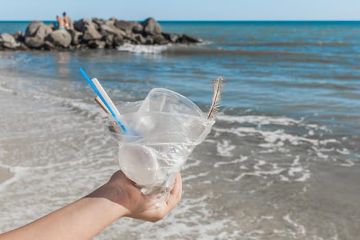 The girl's hand holds a package with a pile of small debris against the background of the beach and the sea. The concept of pollution and ecology. Waste disposal.