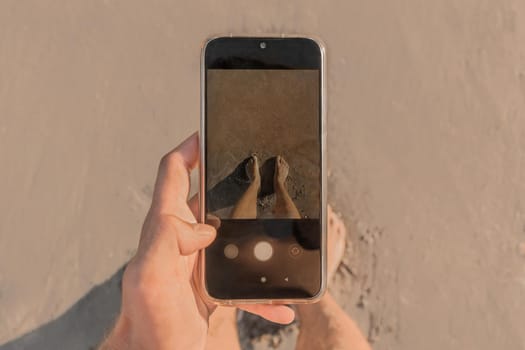 The man's hand holds a mobile phone and takes pictures of his bare feet standing on the wet beach sand by the sea, close up.