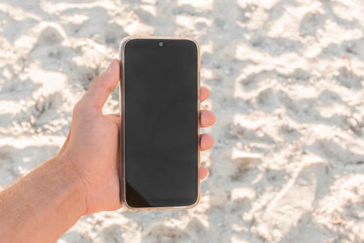 The hand holds a mobile phone with a black screen, a place for text and design, against the background of white beach sand.