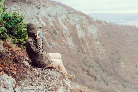 Young woman wearing parka sitting in the mountains and looking into the distance. Space for text in right part of image