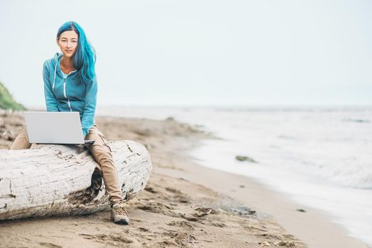 Smiling freelancer girl with blue hair working on laptop on sand beach near the sea, looking at camera. Freelance concept