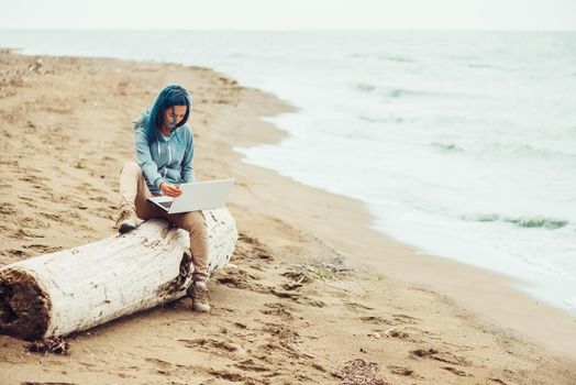 Freelancer young woman with blue hair sitting on tree trunk and working on laptop on sand beach near the sea. Freelance concept