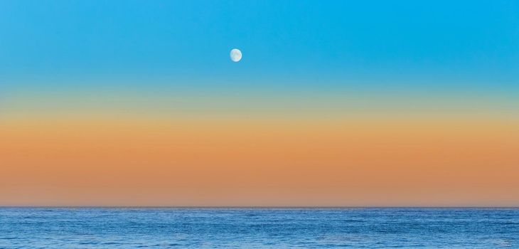 Twilight on the sea beach in the evening with an orange sunset on skyline and blue sky with a moon.