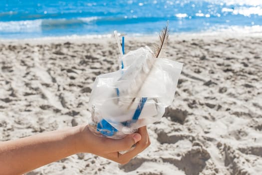 The girl's hand holds a package with a pile of small debris against the background of the beach and the sea. The concept of pollution and ecology. Waste disposal.