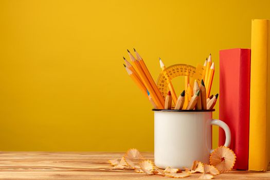 Metal cup with sharp pencils and pencil shavings on wooden desk against yellow background