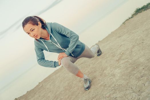 Beautiful sporty girl stretching her legs and preparing to run on coast in summer in the morning, workout. Concept of healthy lifestyle