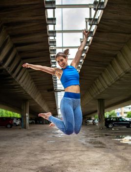 Athletic woman smiling happy exercising outdoors in parking lot. Sporty girl jumping with arms up and legs bent.