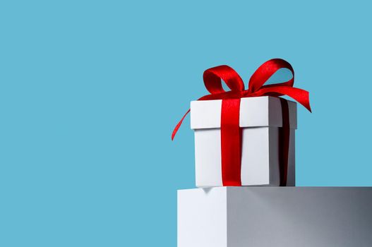 Creative Christmas Concept. Close-up White Gift Box with Red Ribbon on Stand on Blue Background.