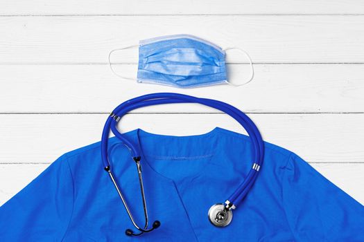 Doctor medical uniform with stethoscope and face mask on white wooden background close up