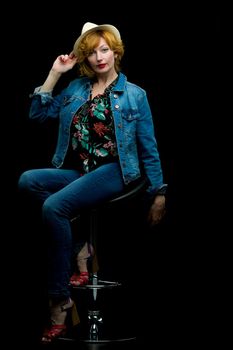 Beautiful young woman posing in a studio studio on a bar stool holding a hat in her hand. The concept of style and fashion. Isolated on black background.