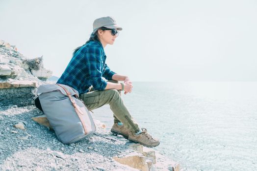 Beautiful traveler young woman sitting on stone coast with backpack and enjoying view of sea in summer