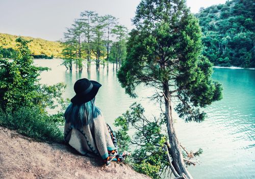 Young woman in hat and poncho resting on the shore of lake and enjoying view of nature