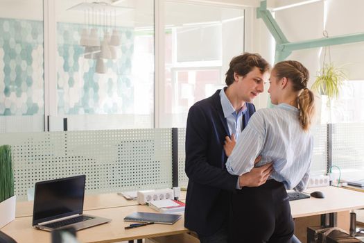 Two young people embracing and looking at each other while flirting near desk in modern office