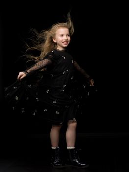 A strong wind blows on the face and clothes of a beautiful little girl of school age. Hair and clothes are fluttering in the wind, and the girl is happy about it. On a black background. Studio photo session