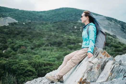 Beautiful traveler young woman sitting on peak of cliff and enjoying view of mountains in summer outdoor