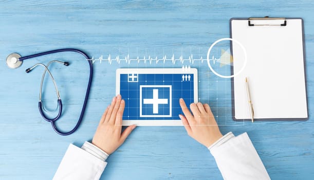 Top view of doctor hands with tablet computer. Therapist in white coat sitting at blue wooden desk. Examination and consultation in clinic. Medical application and online healthcare services.