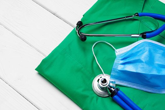 Doctor medical uniform with stethoscope and face mask on white wooden background close up